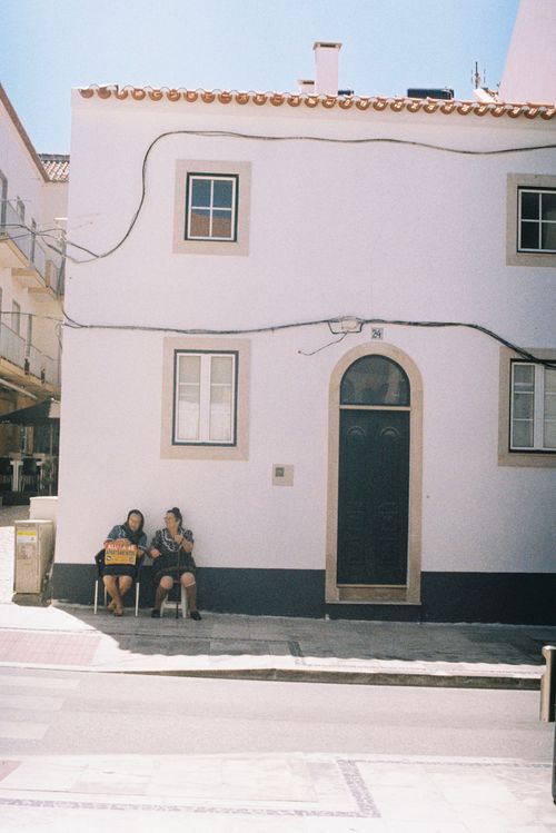 Free Charming street view in Nazaré, Portugal featuring two residents enjoying a sunny day by a traditional house. Stock Photo