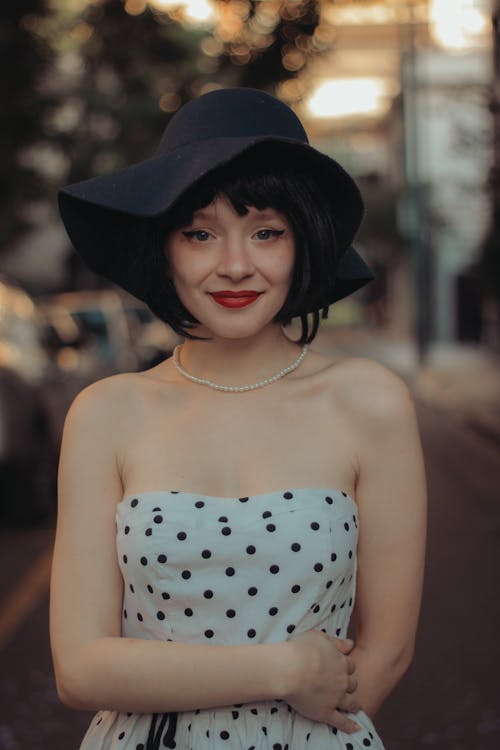 Smiling Model in a White Off-the-Shoulder Dress with Polka Dots and Black Wide Brim Hat on the Street