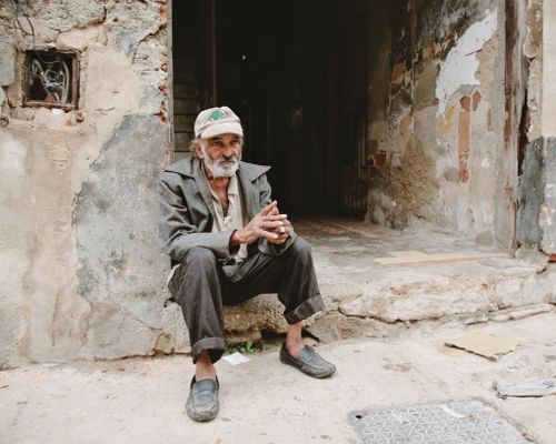 Free An elderly man with a beard sits on worn steps in a decaying urban environment. Stock Photo