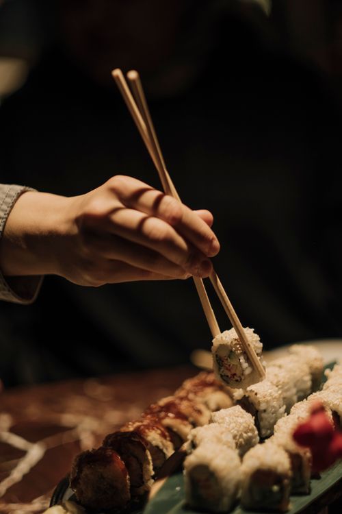 Free Close-up of a hand using chopsticks to pick sushi roll on a plate in a dimly lit setting. Stock Photo