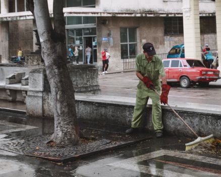 A city worker cleans a wet urban street with a broom, surrounded by pedestrians and a parked car.