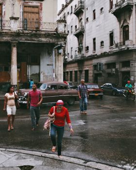Rainy urban scene with people walking past vintage cars in a historic city area.