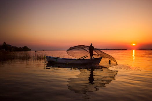 A fisherman casts his net from a boat on a tranquil lake at sunrise, reflecting vibrant orange hues.