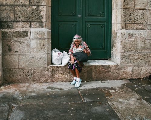 Free An elderly woman peacefully sitting by a large green door, highlighting urban solitude. Stock Photo