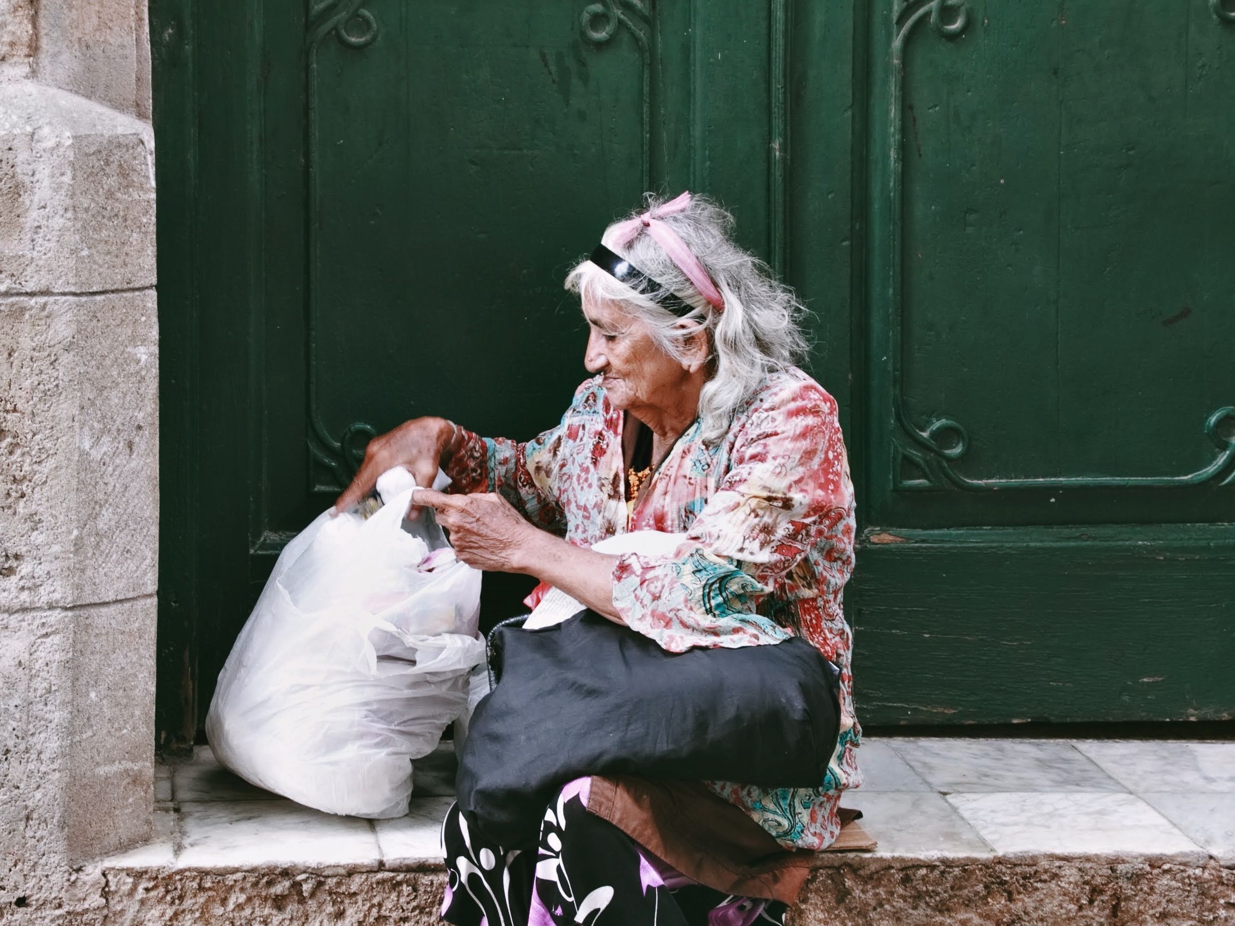 Elderly woman sitting by a green door with bags in an urban setting.
