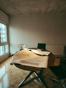 A modern office with architectural blueprints on a wooden round table by the window.