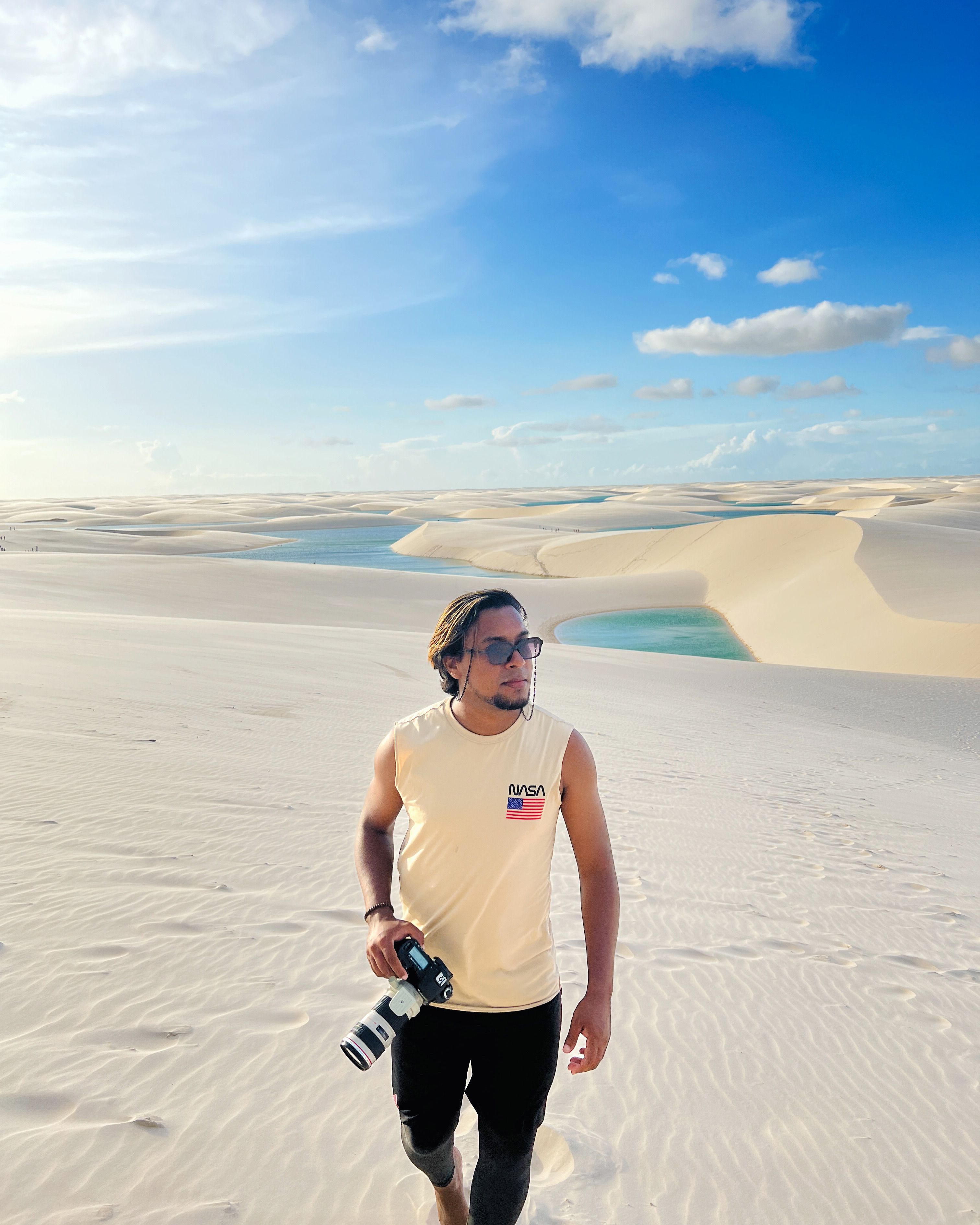 A man with a camera walks through sunlit sand dunes under a clear blue sky.