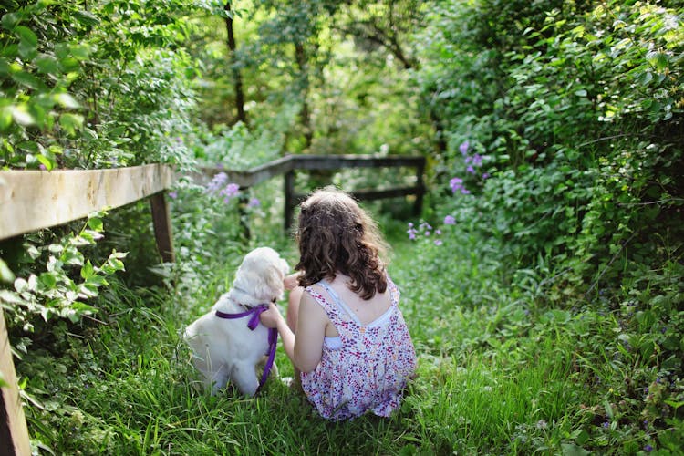 Girl And Puppy Sitting On Green Grass Surrounded With Plants During Daytime