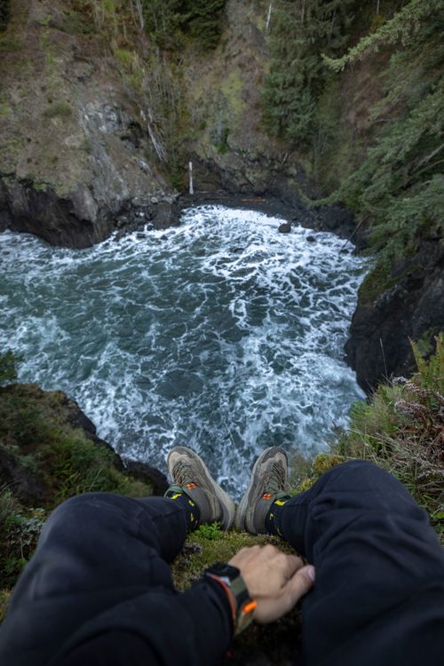Free A daring adventurer sits with legs dangling over a rugged cliff edge, overlooking churning waters below. Stock Photo
