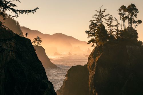 Free Silhouetted figure stands on a rocky coastline at sunset, with misty seascape and trees. Stock Photo