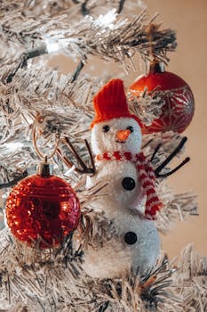 A cozy holiday scene with a snowman ornament and red baubles on a frosted Christmas tree.