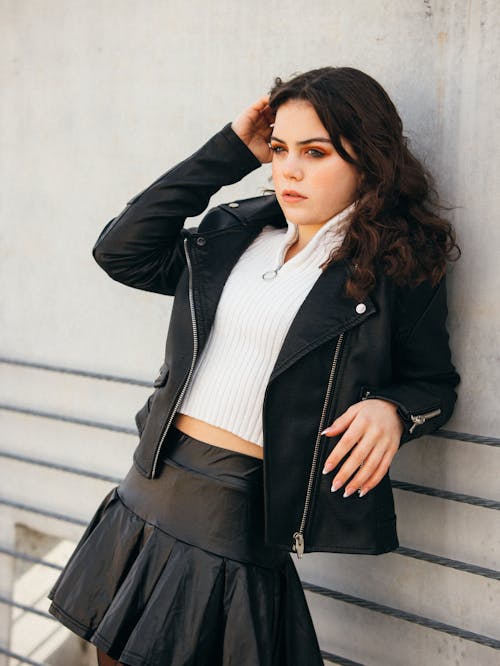 A woman in a skirt and leather jacket leaning against a wall