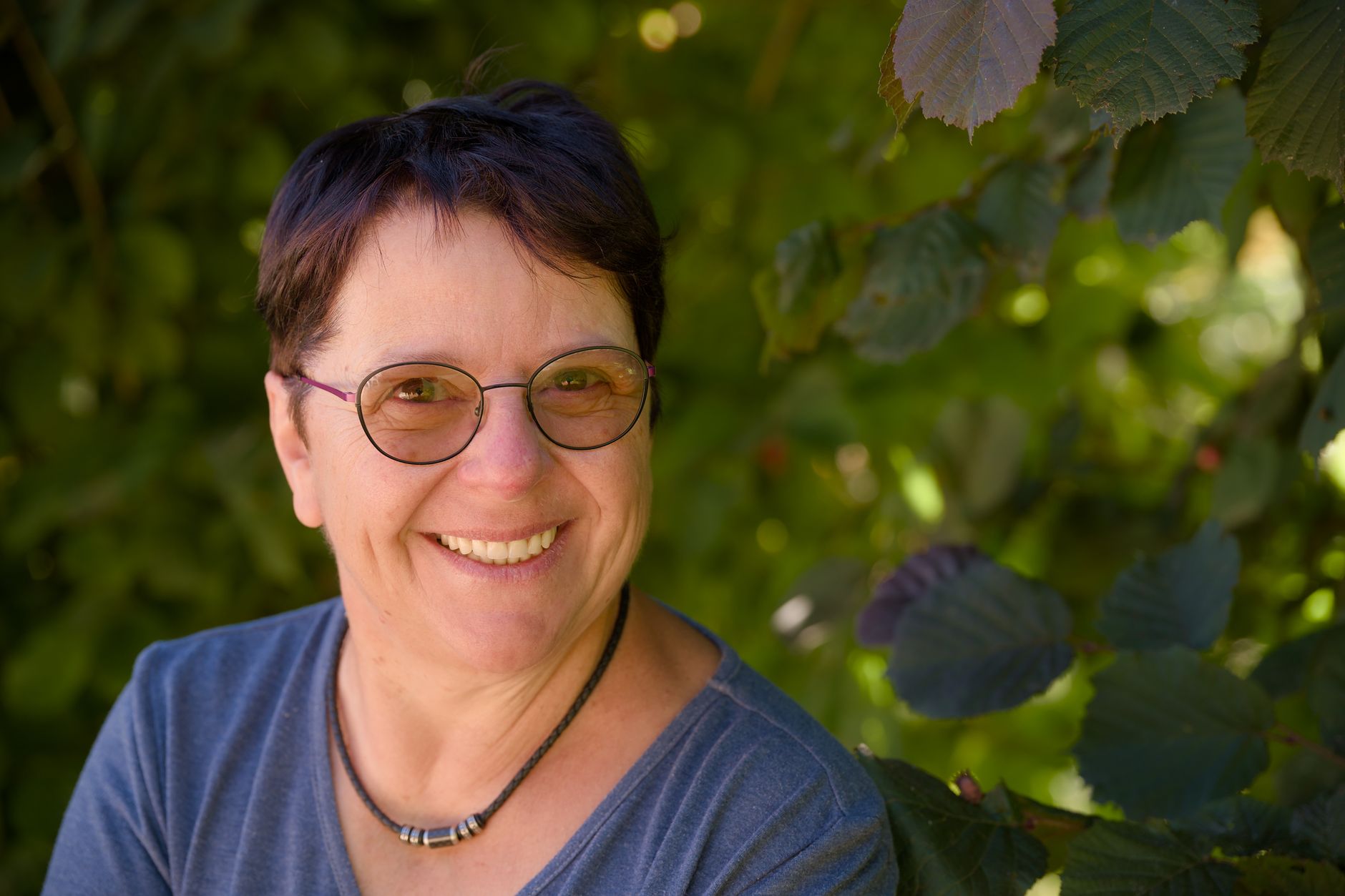 Portrait of a cheerful woman wearing eyeglasses, smiling amidst summer foliage.