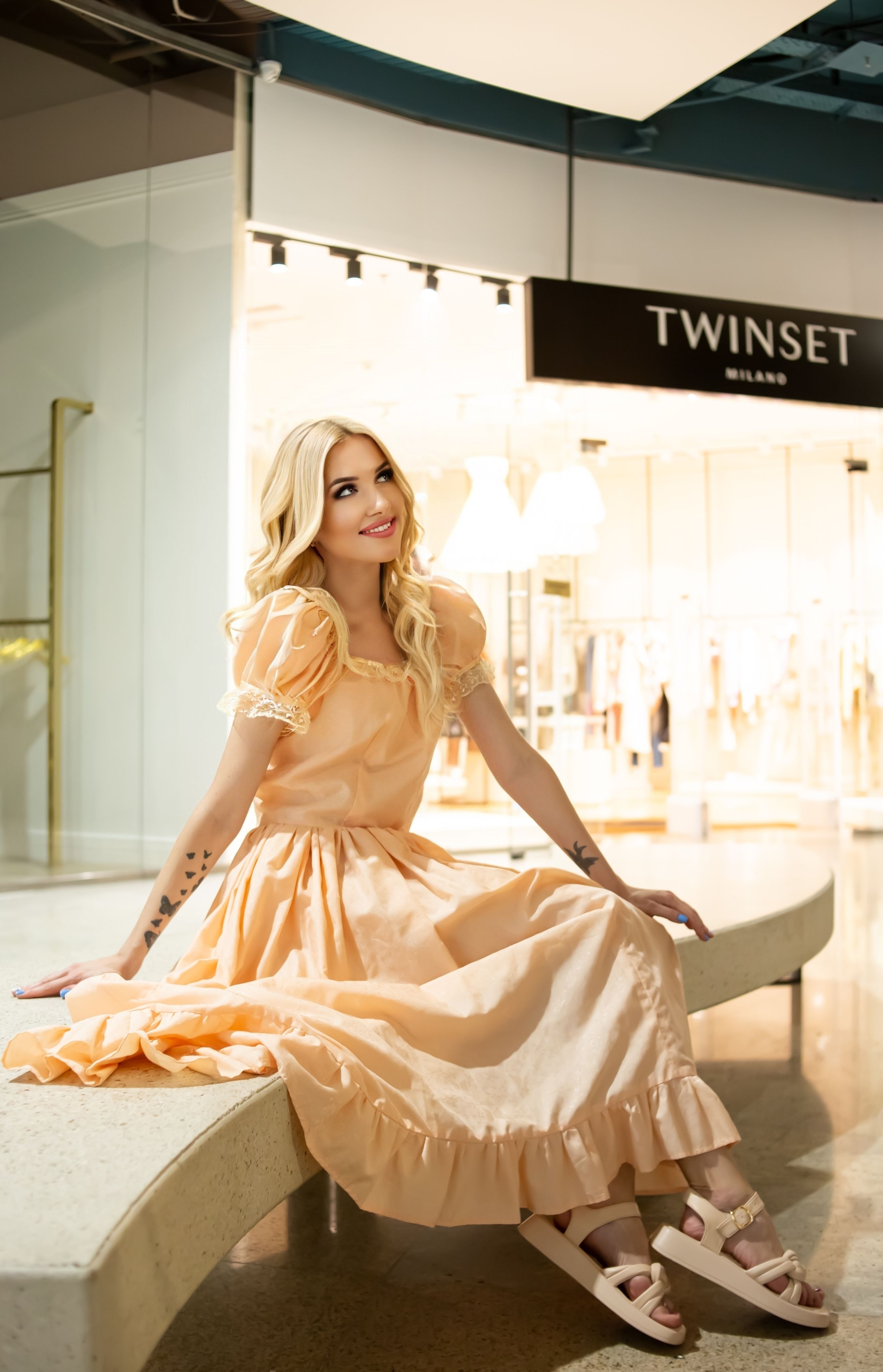 Woman in a peach dress sitting inside a shopping mall, exuding elegance and fashion.