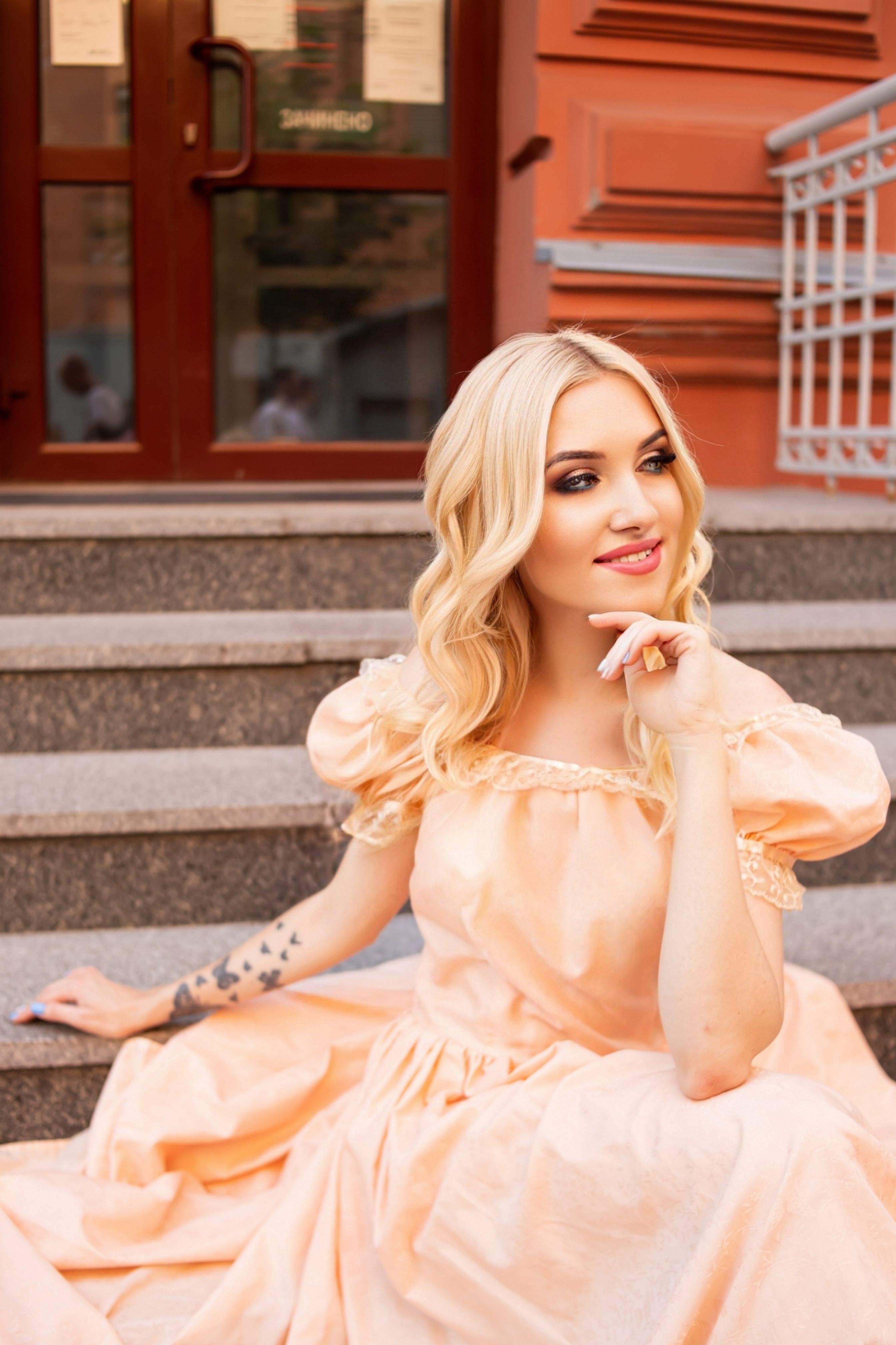 Stylish woman in a peach gown sitting on outdoor steps, exuding elegance and charm.