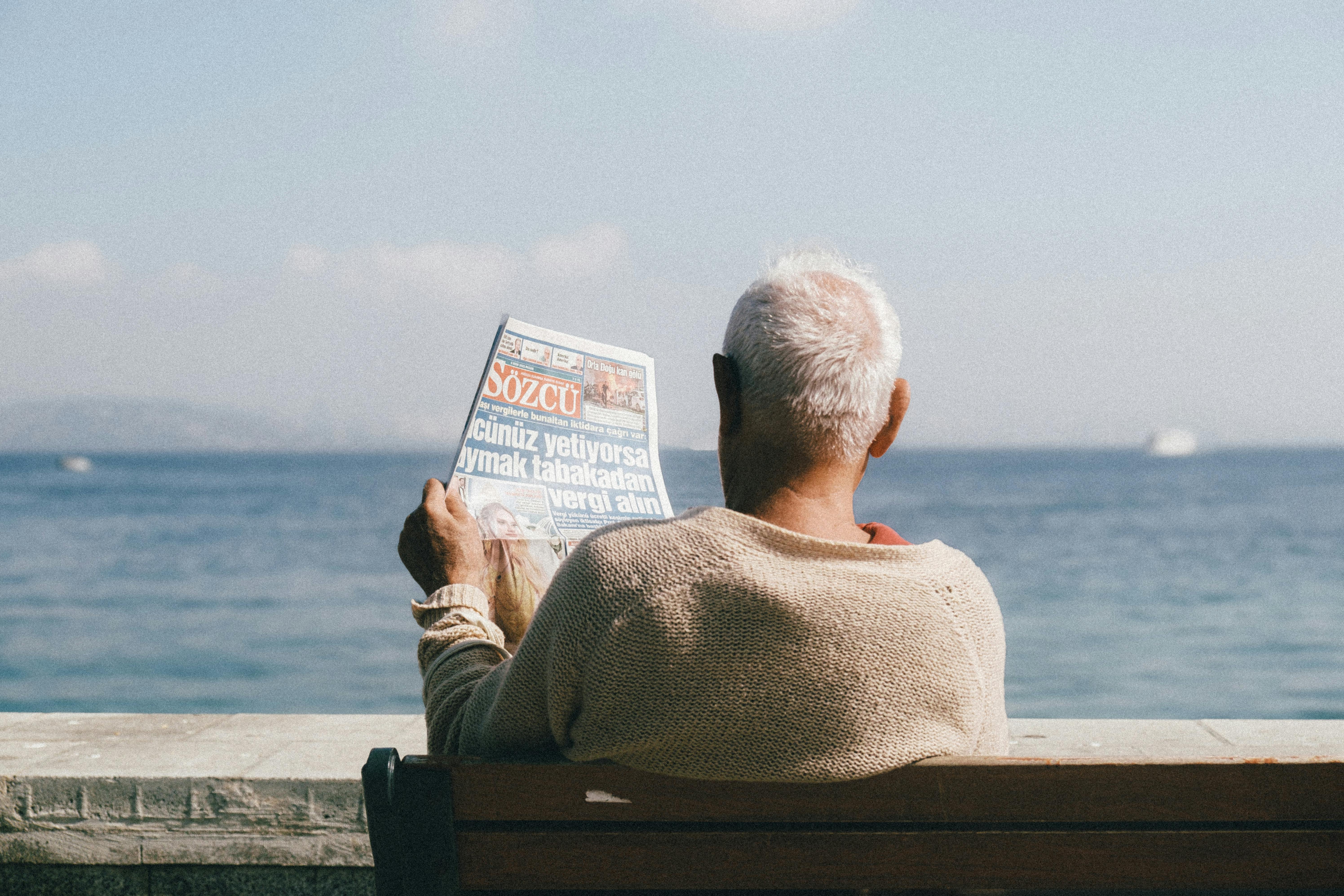 pensioner reading newspaper on bench by sea