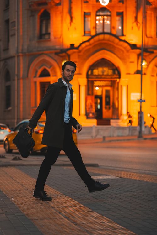 Free A fashionable man in a suit and coat crossing a city street at night with a lit building backdrop. Stock Photo