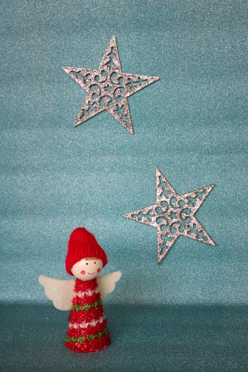 A cute Christmas angel figurine with white wings and a red hat against a blurred glittering pastel blue background with two shining silver stars. Vertical orientation with empty space for ...