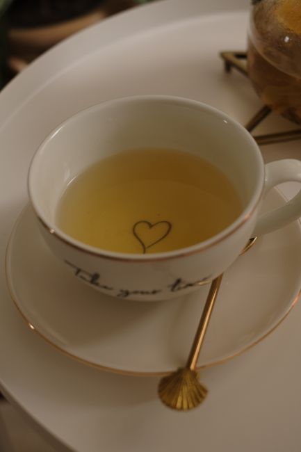 A close-up of a porcelain tea cup with a heart detail on a golden spoon and tray.