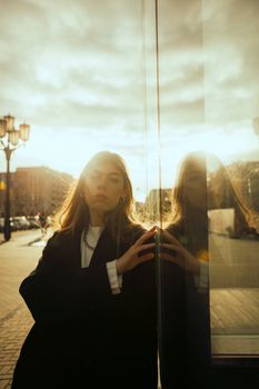 A young woman poses beside a glass window, capturing a sunset reflection in an urban setting.