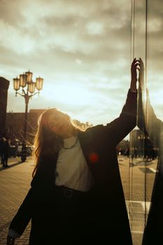 Elegant woman in coat poses against reflective surface with sunset backdrop, creating a dramatic silhouette.