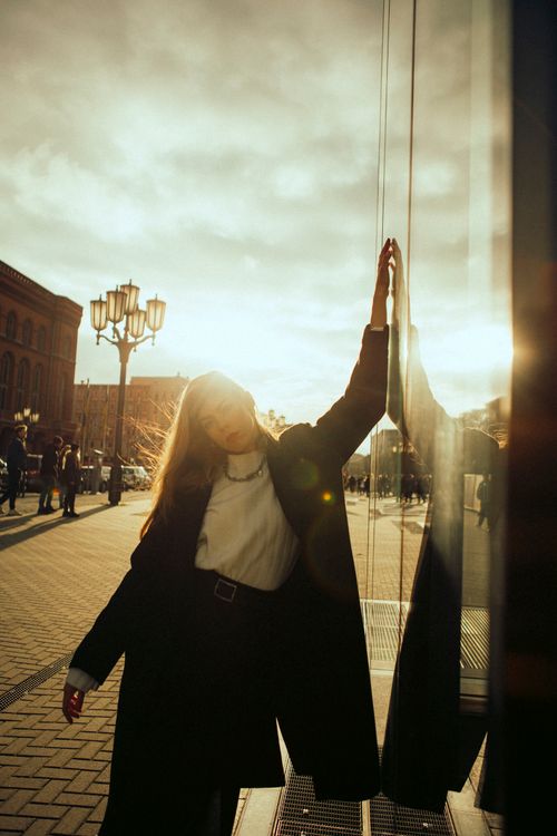 Free Chic woman in coat poses elegantly by a reflective wall during sunset. Fashion and urban vibes. Stock Photo