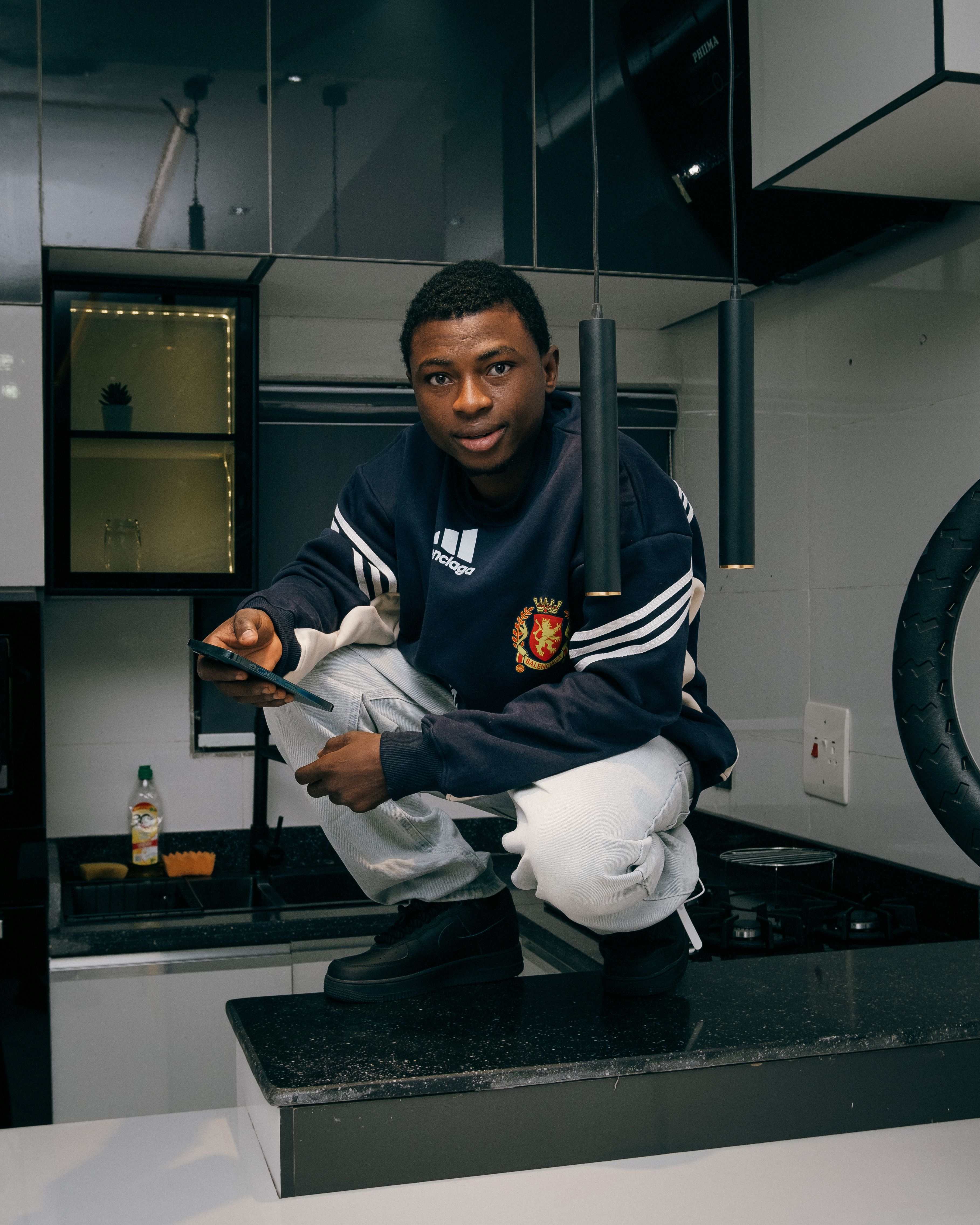 Young man using smartphone while squatting on a kitchen countertop. Modern interior with appliances visible.