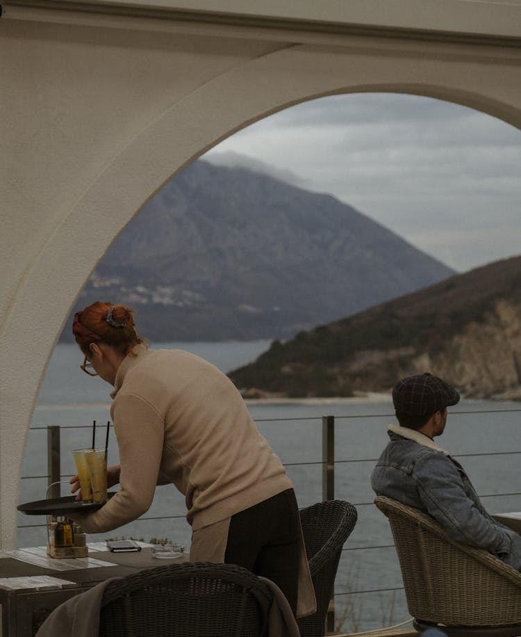 Waitress Cleaning A Table On The Restaurant Terrace Overlooking The Bay And A Customer Enjoying The View