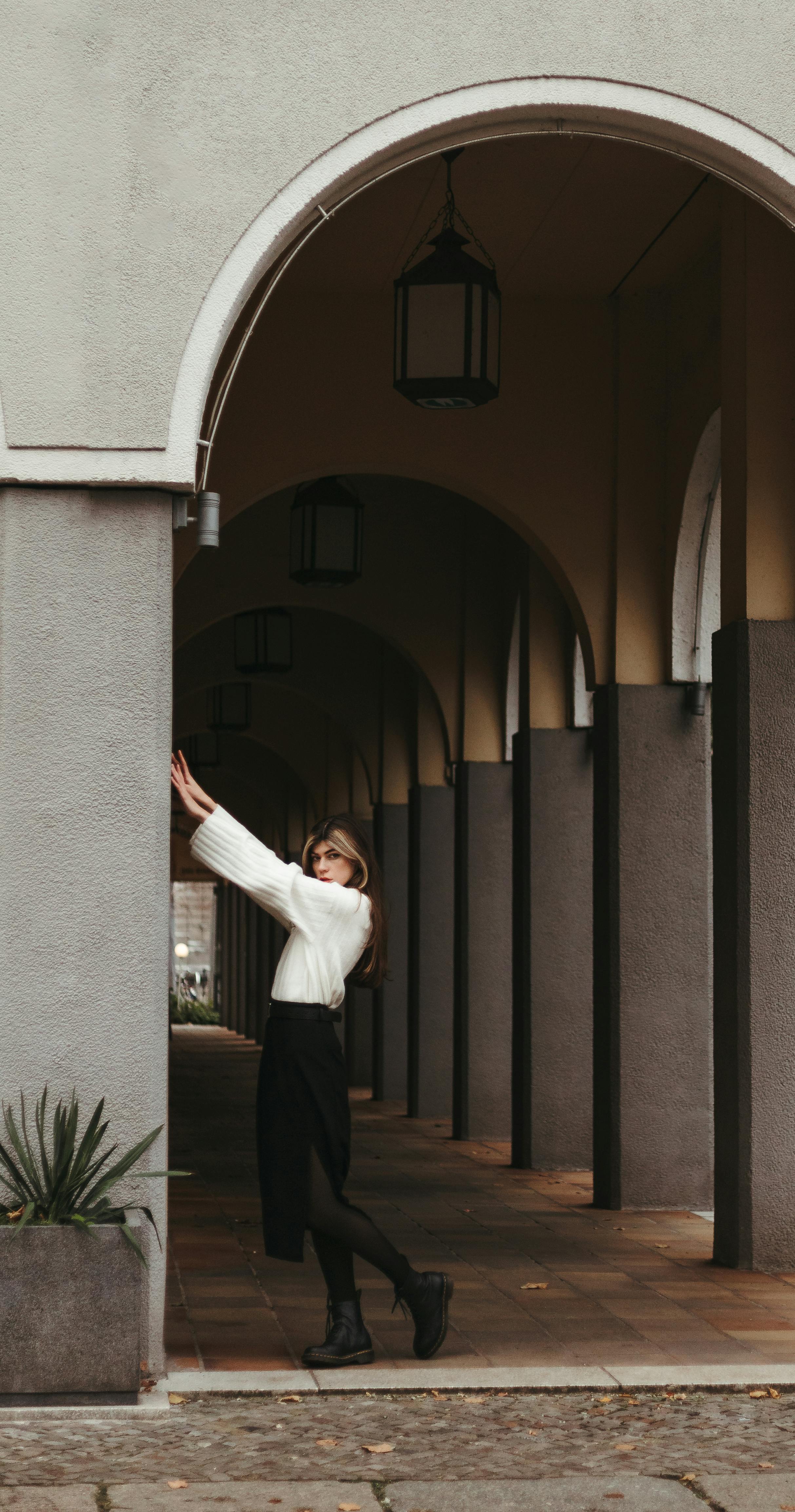 Elegant woman posing under a series of arches in an urban setting, showcasing fashion and architecture.
