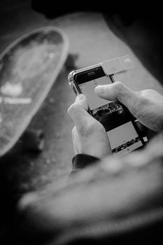 Black and white photo of person holding smartphone next to a skateboard on city street.