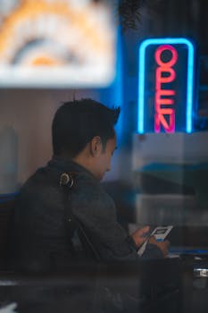 Young man sits in a cafe checking his phone under a neon open sign.