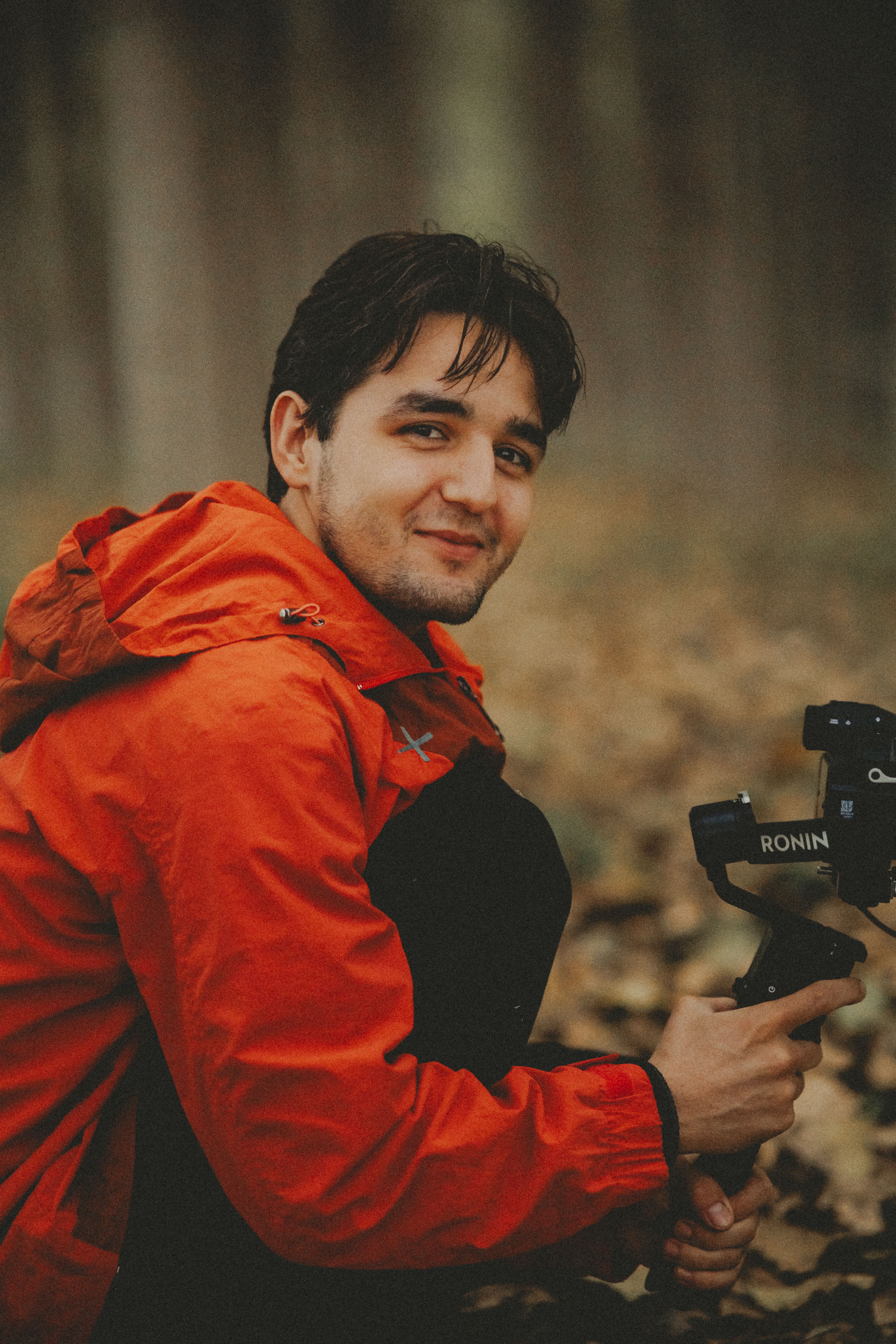 Portrait of a young man in a red jacket holding a camera in a forest setting, smiling.