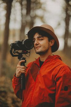 Man in a red jacket and hat holding a camera outdoors in a wooded area.