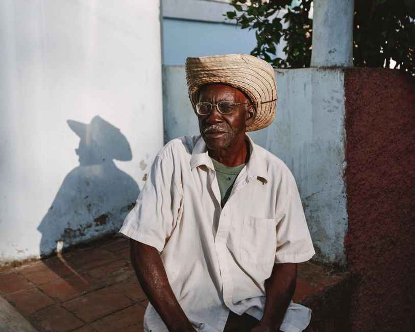Portrait of an elderly man sitting outside, wearing eyeglasses and a straw hat.