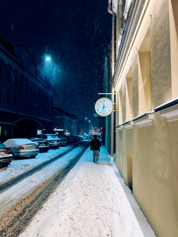 Person Walking In Snow Near Street And Parked Cars At Night