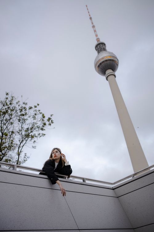 Free A young woman stands near a railing with the Berlin TV Tower in the background under an overcast sky. Stock Photo