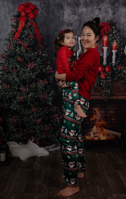 Smiling Woman in Pajamas with Her Little Daughter in Her Arms Near a Christmas Tree and Ornaments