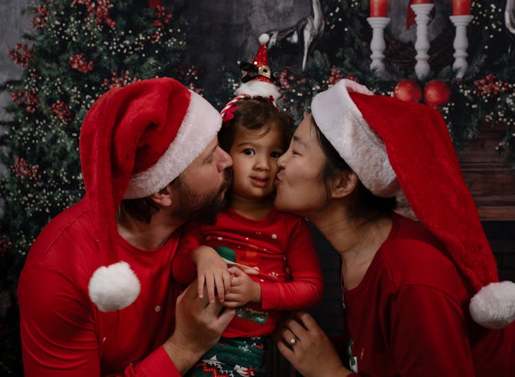 Parents Kissing Their Little Daughter On The Cheeks Dressed In Christmas Pajamas And Santa Hats