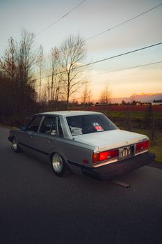 A classic modified Toyota car with a lowered stance parked on a rural road at sunset.