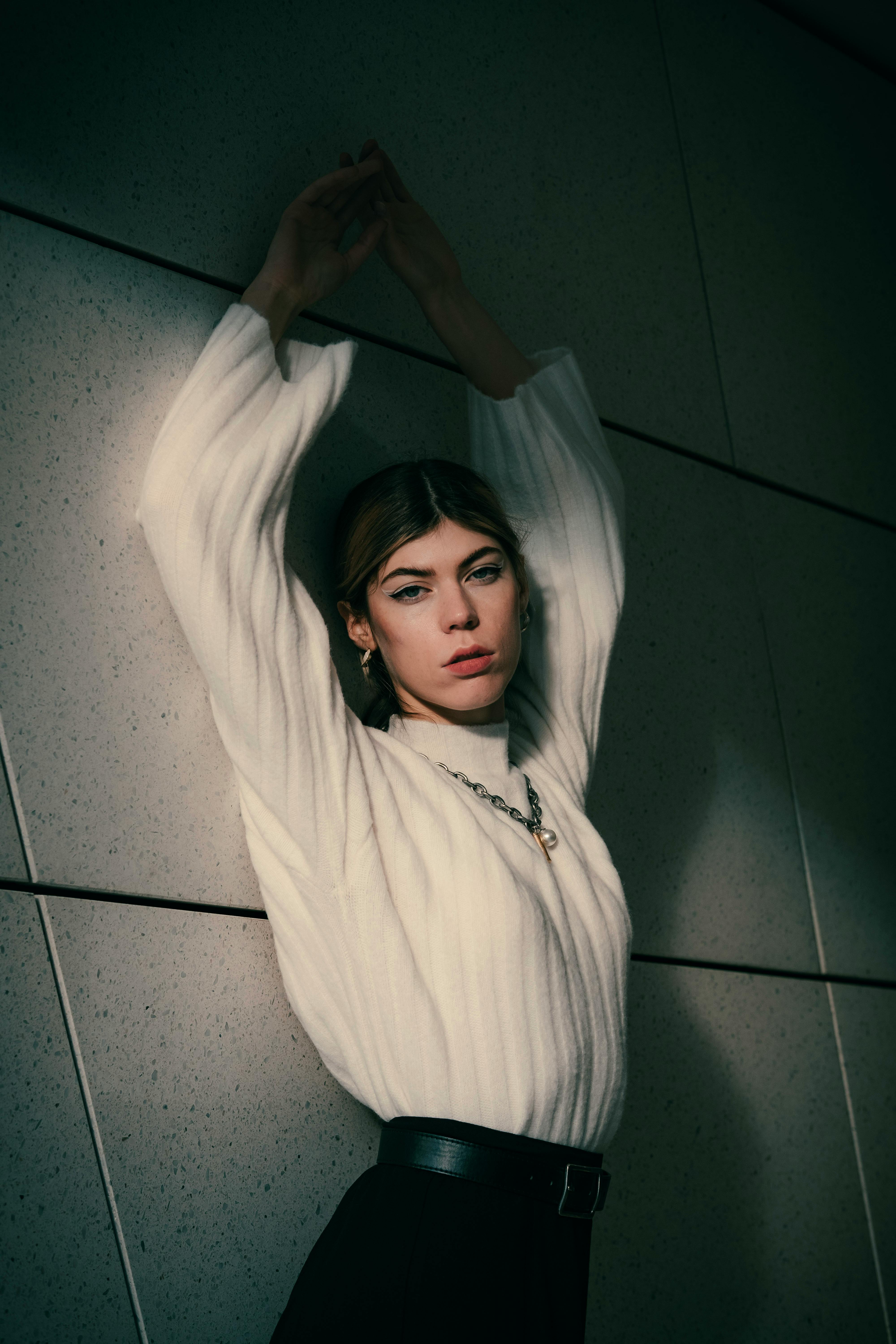 Model posing with arms raised against a tiled wall, wearing a stylish white sweater.