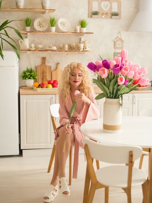 Free Blonde woman in a pink robe surrounded by vibrant tulips in a bright kitchen, exuding elegance and relaxation. Stock Photo