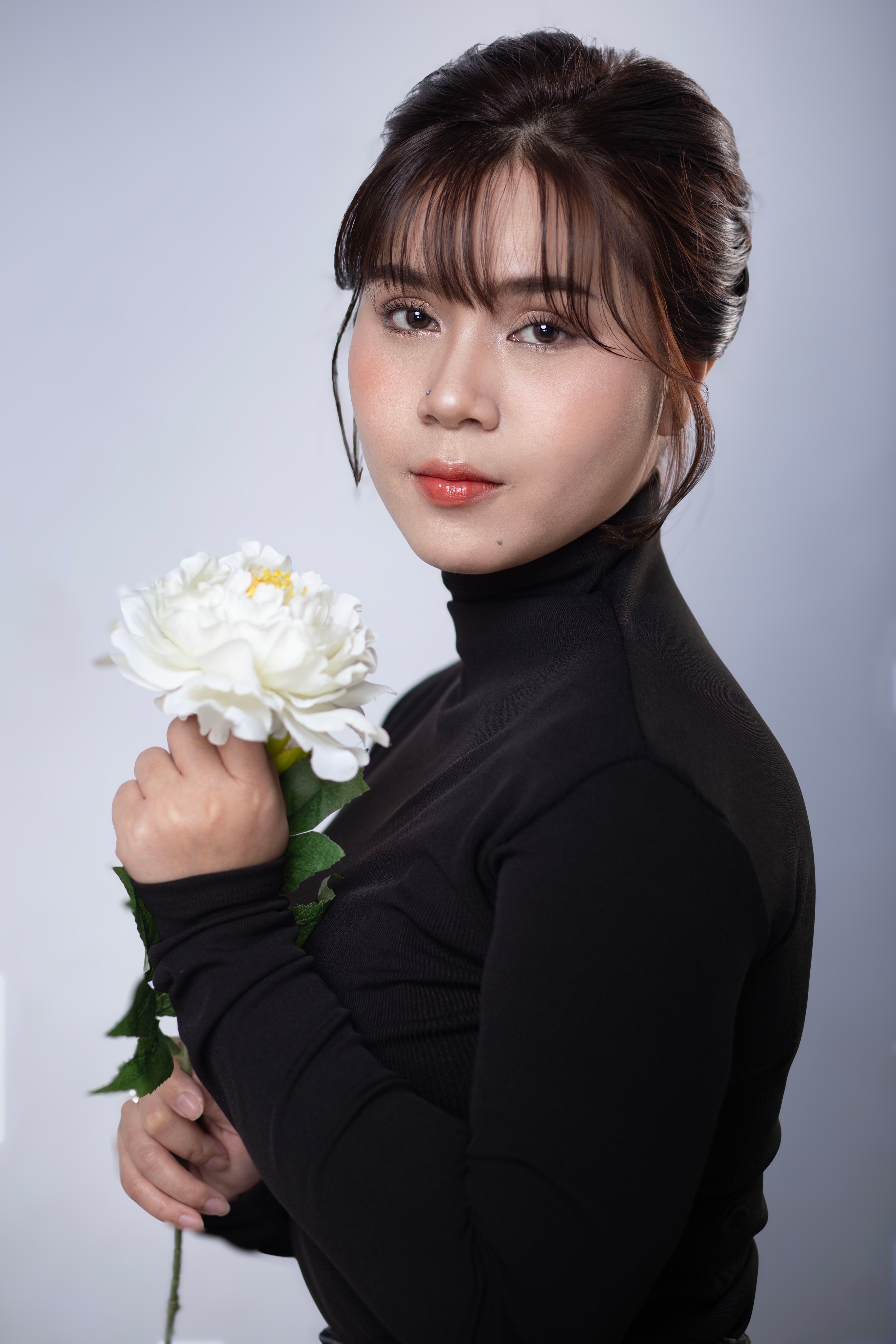 Striking portrait of a woman in black turtleneck holding a white flower against a neutral background.