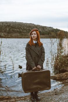 A redhead woman in a coat holds luggage by a tranquil lake in Ankara, Türkiye.