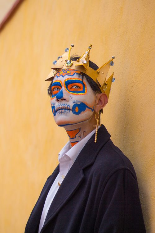 Free Man with sugar skull face paint and crown stands against a yellow wall for Dia de los Muertos celebration. Stock Photo