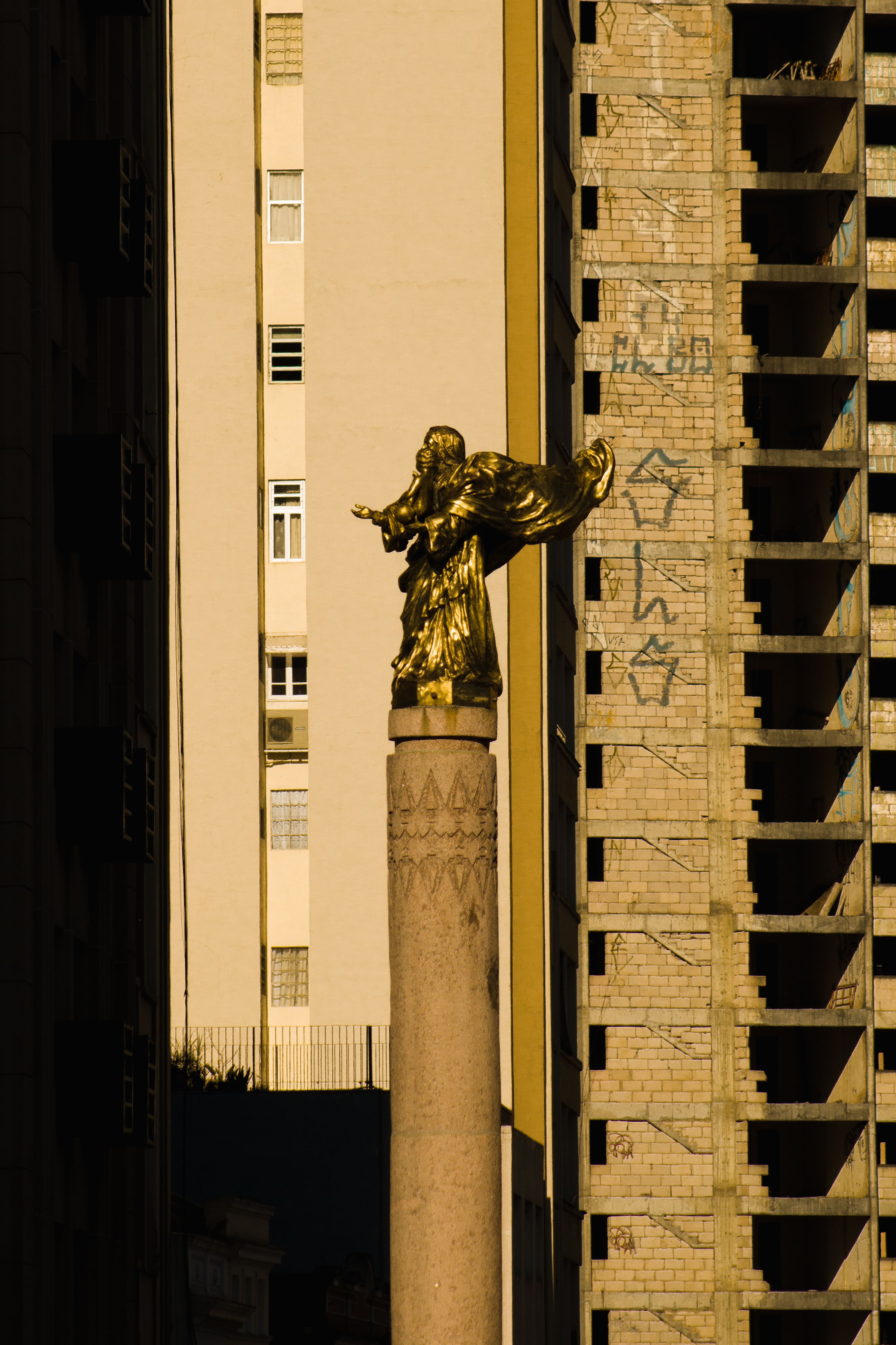 Golden angel statue on a column with urban buildings in the background, captured at sunset.