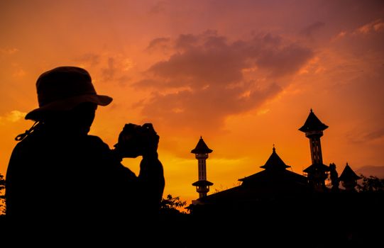 Silhouette of a photographer capturing a mosque at sunset with an orange sky.