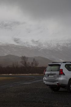 White off-road SUV parked on gravel path with snowy mountain backdrop under cloudy skies.