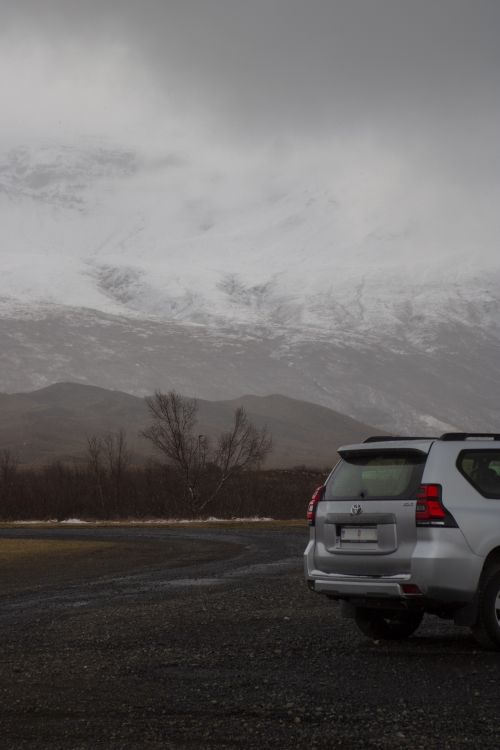 Free White off-road SUV parked on gravel path with snowy mountain backdrop under cloudy skies. Stock Photo