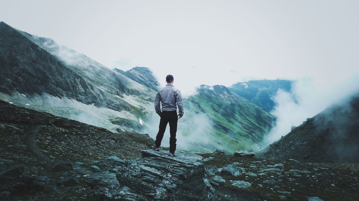 A man in a jacket stands on a rocky mountain peak surrounded by clouds and mist, capturing a sense of adventure.