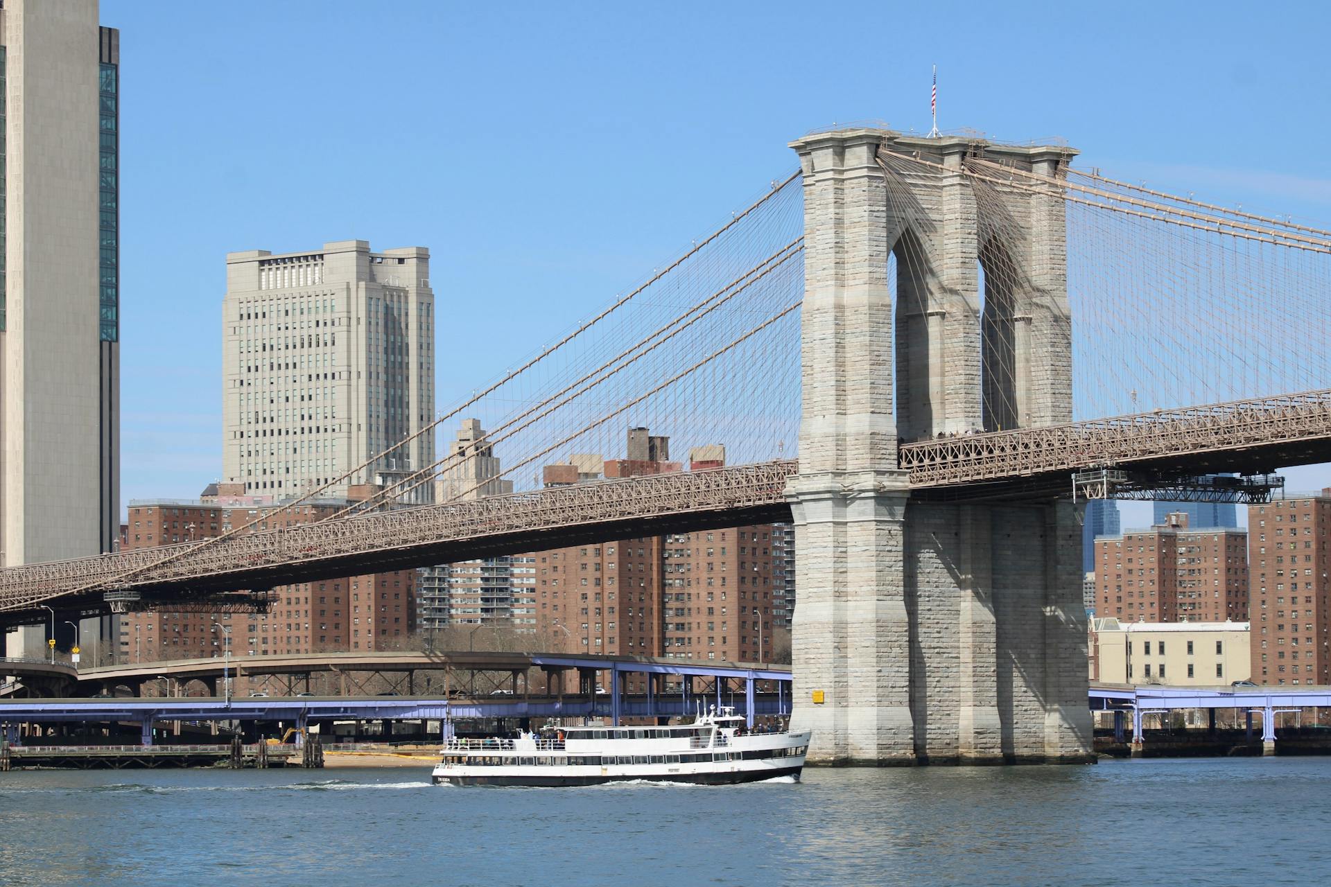 Scenic view of Brooklyn Bridge with a boat on the East River and New York City's skyline in the background.